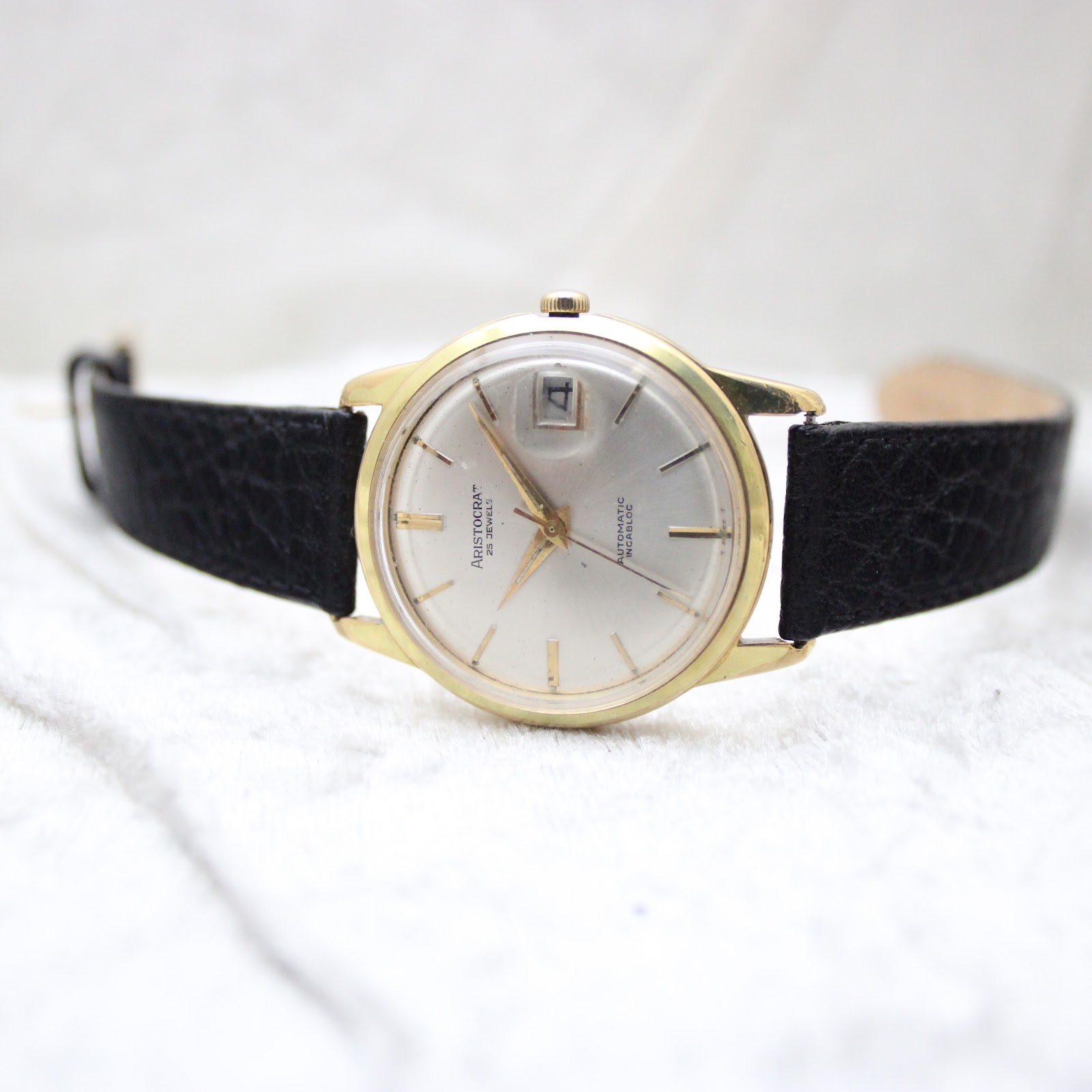 Top-down front view of the Aristocrat watch on a white textured surface, showing the dial and black strap laid flat.