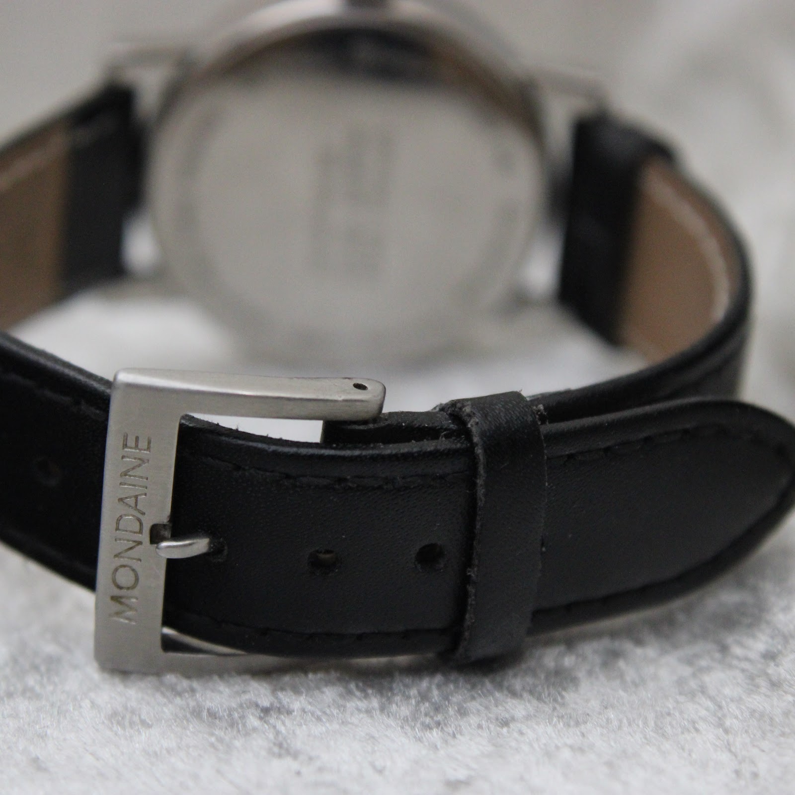 Close-up shot of the black leather strap with a brushed stainless steel buckle, clearly showing the "MONDAINE" signature engraved on it.