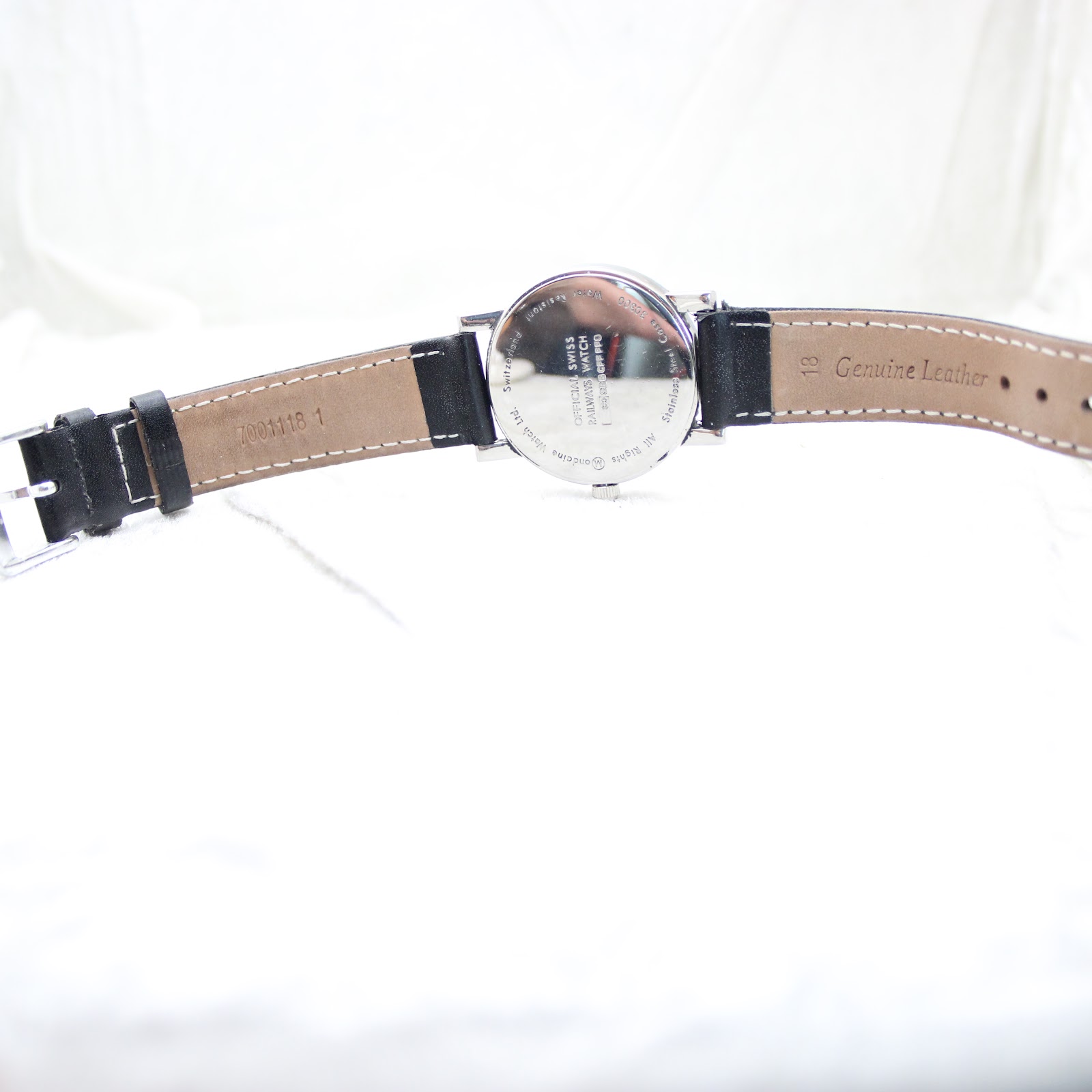 Overhead view of the watch from the back, displaying the black leather strap unfolded and the stainless steel case back with its various engravings.