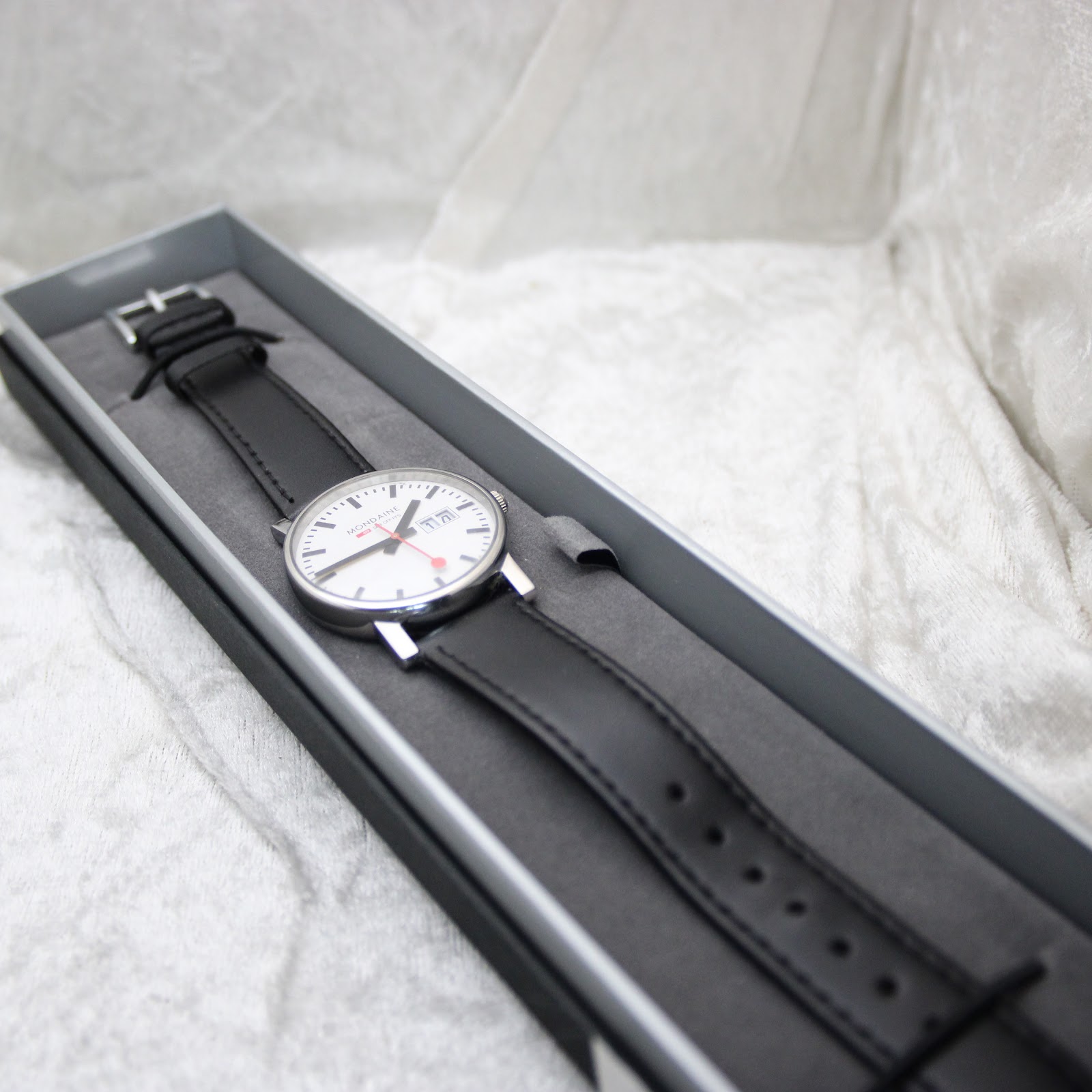 The Mondaine watch resting inside a simple, long grey presentation box, with its black leather strap laid out, suggesting careful storage.