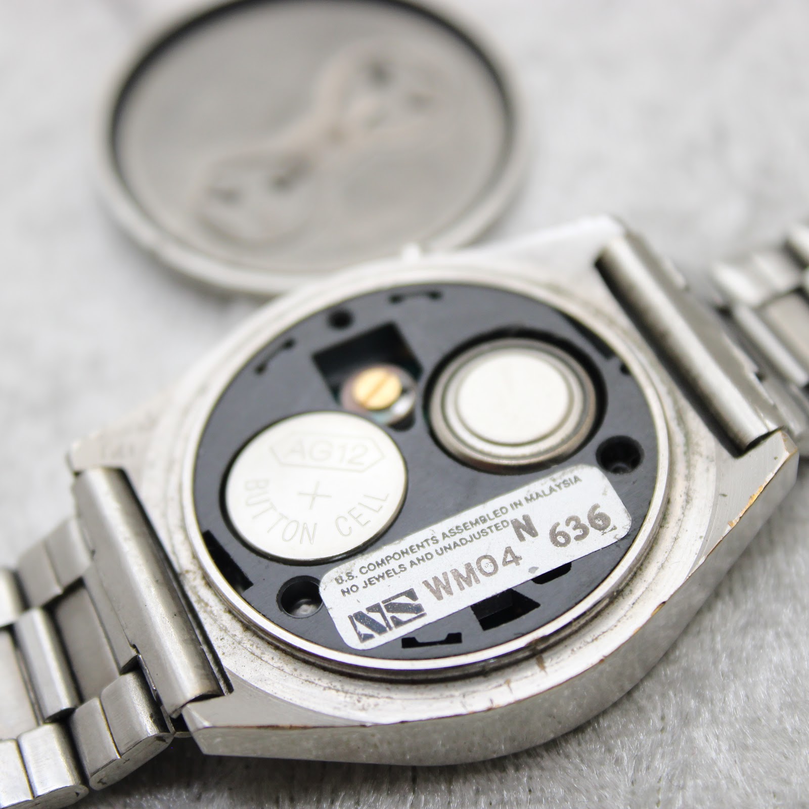 Interior view of the watch movement with the case back removed, showing two AG12 button cell batteries and a "US components assembled in Malaysia" sticker.