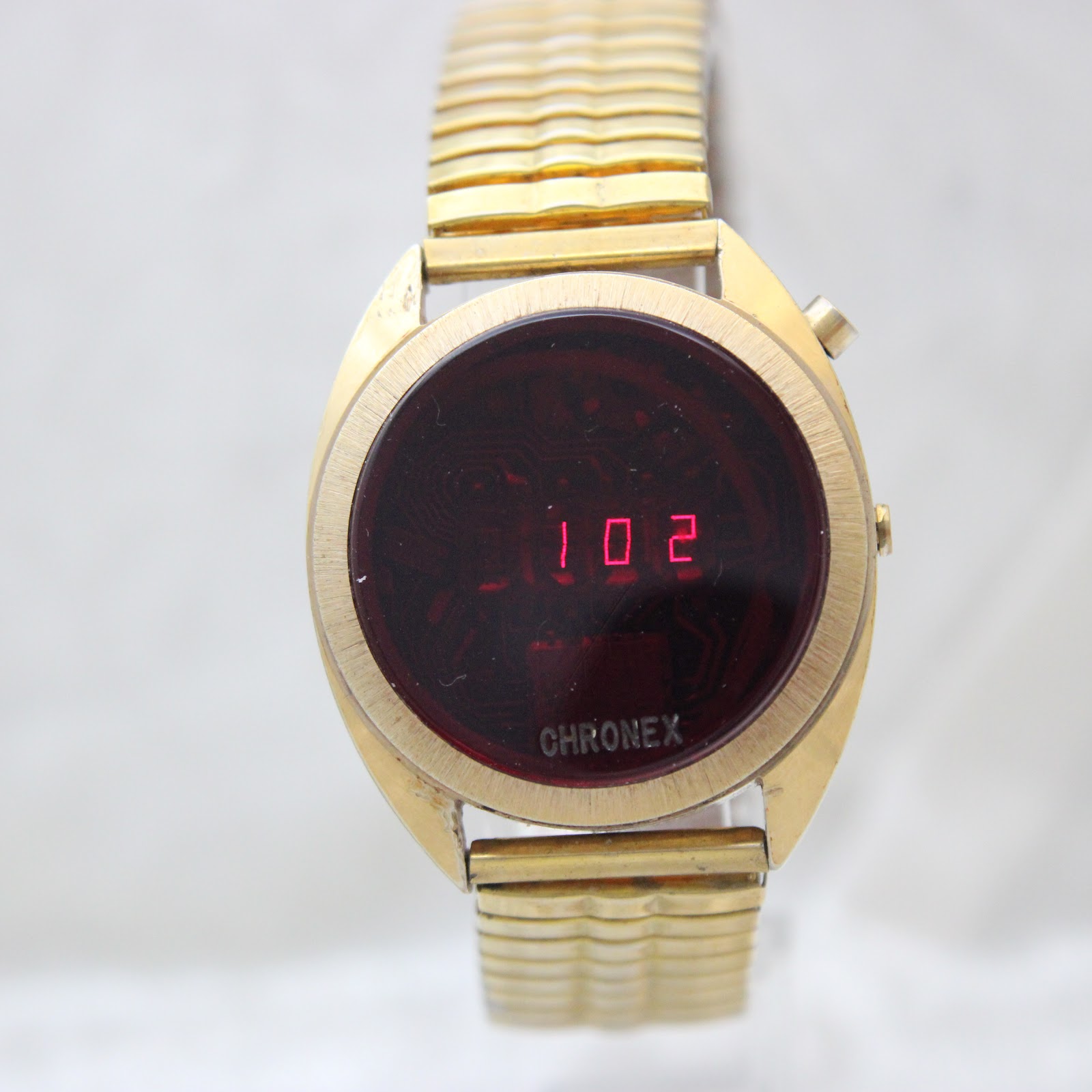 Front view of the Chronex LED watch displaying "1 0 2" in red, with 'CHRONEX' visible below the display, all within a gold-tone case and expansion bracelet.