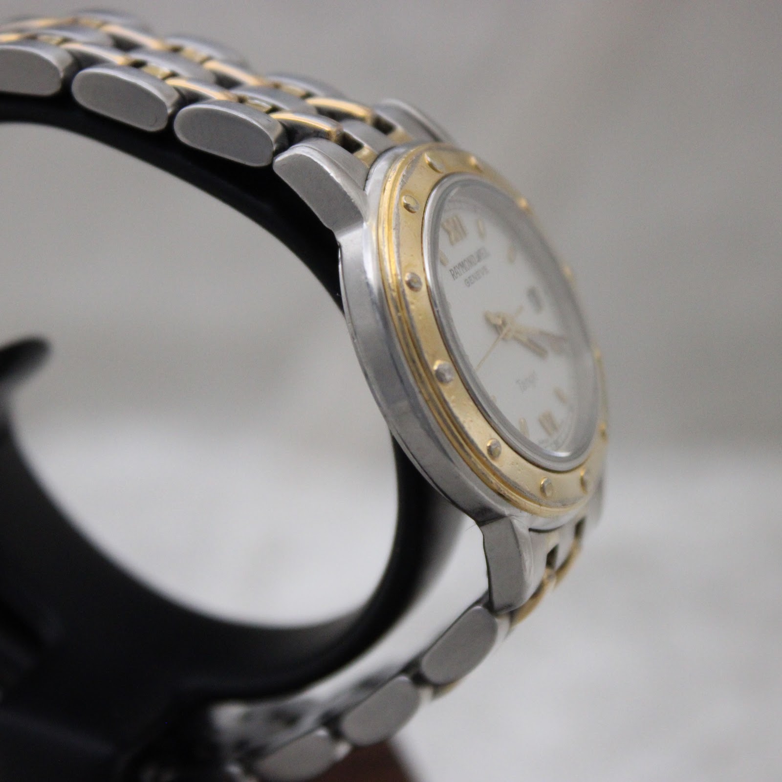 Angled side view of the Raymond Weil Tango watch, highlighting the stainless steel case and gold bezel.