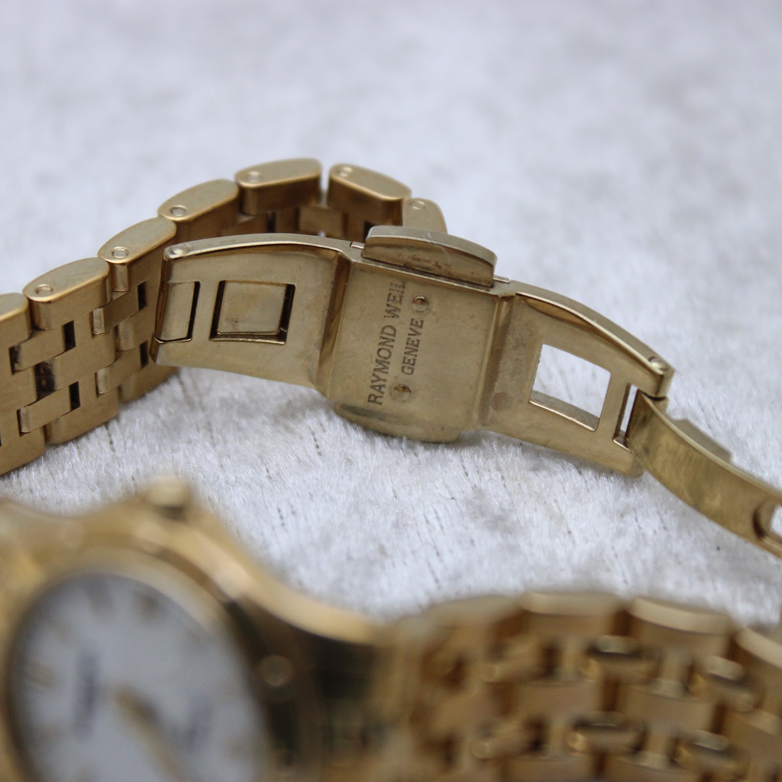 Close-up of the watch's gold-tone deployment clasp, clearly showing the 'Raymond Weil Geneve' signature engraving.