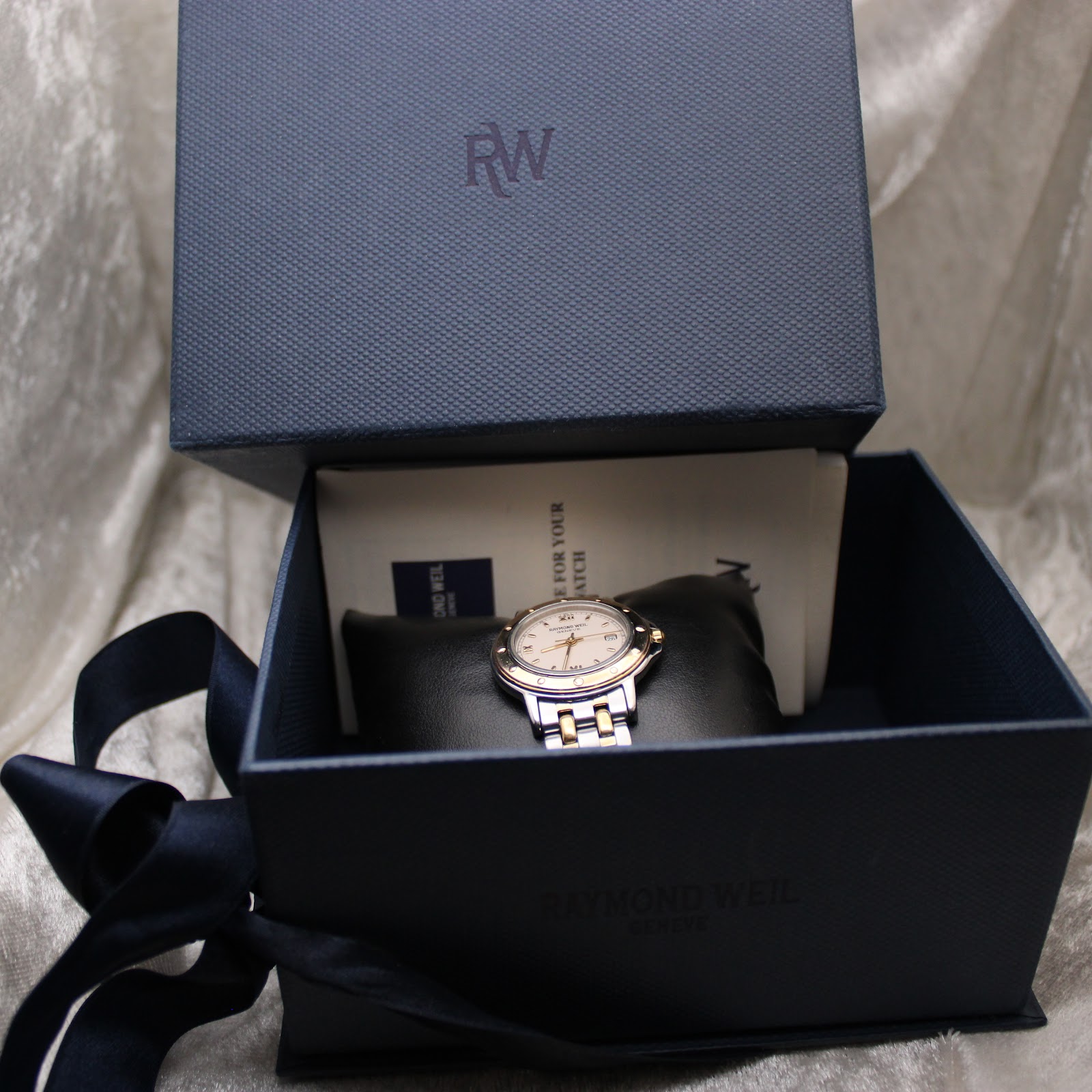 Overhead view of the Raymond Weil watch inside its original dark blue textured box with 'RW' logo, along with papers and cushion.