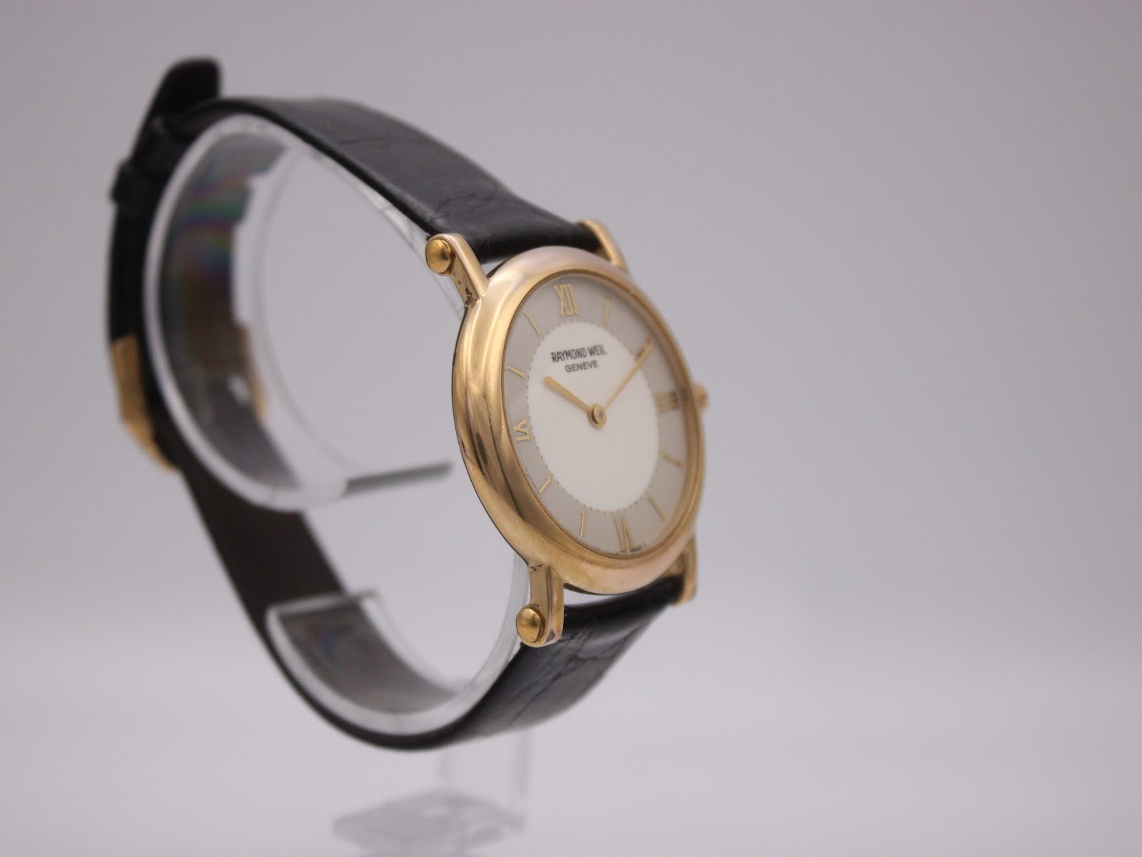 Angled left side view of the Raymond Weil watch, highlighting the gold-plated crown and case profile on a clear display stand.