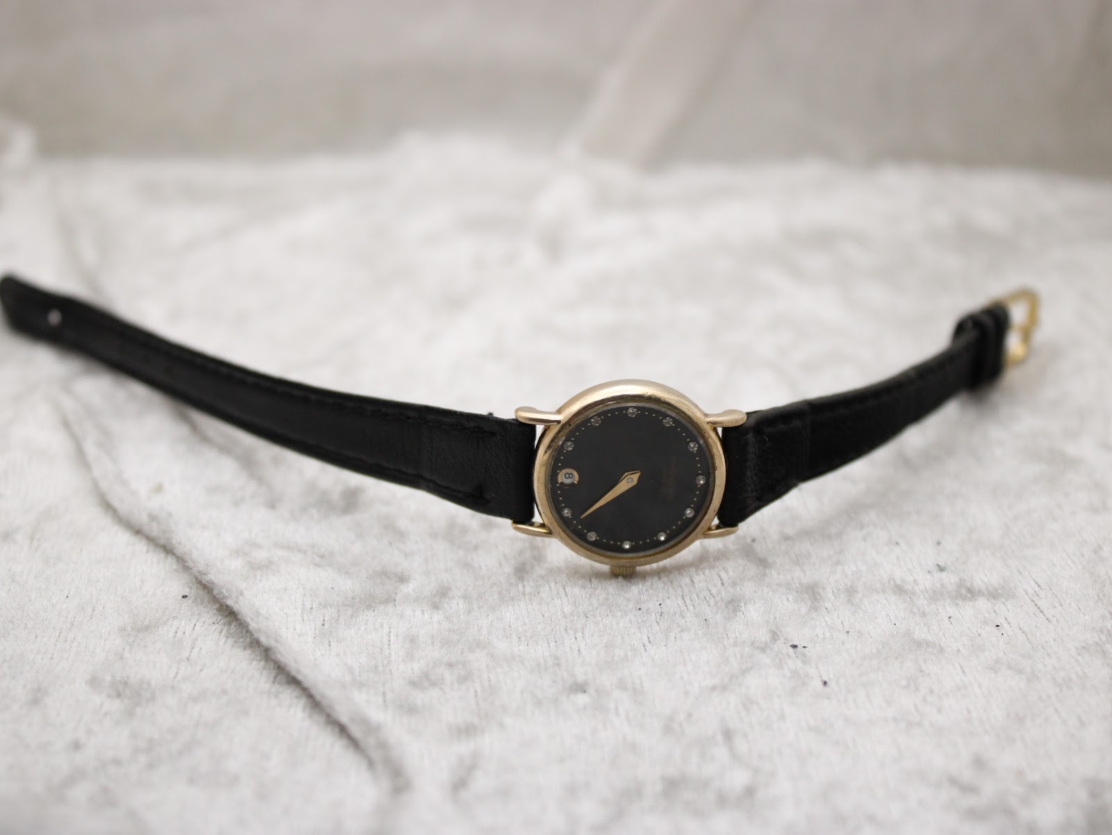 Top-down view of the Raymond Weil watch with a black dial and gold case, laid flat on a white textured surface.
