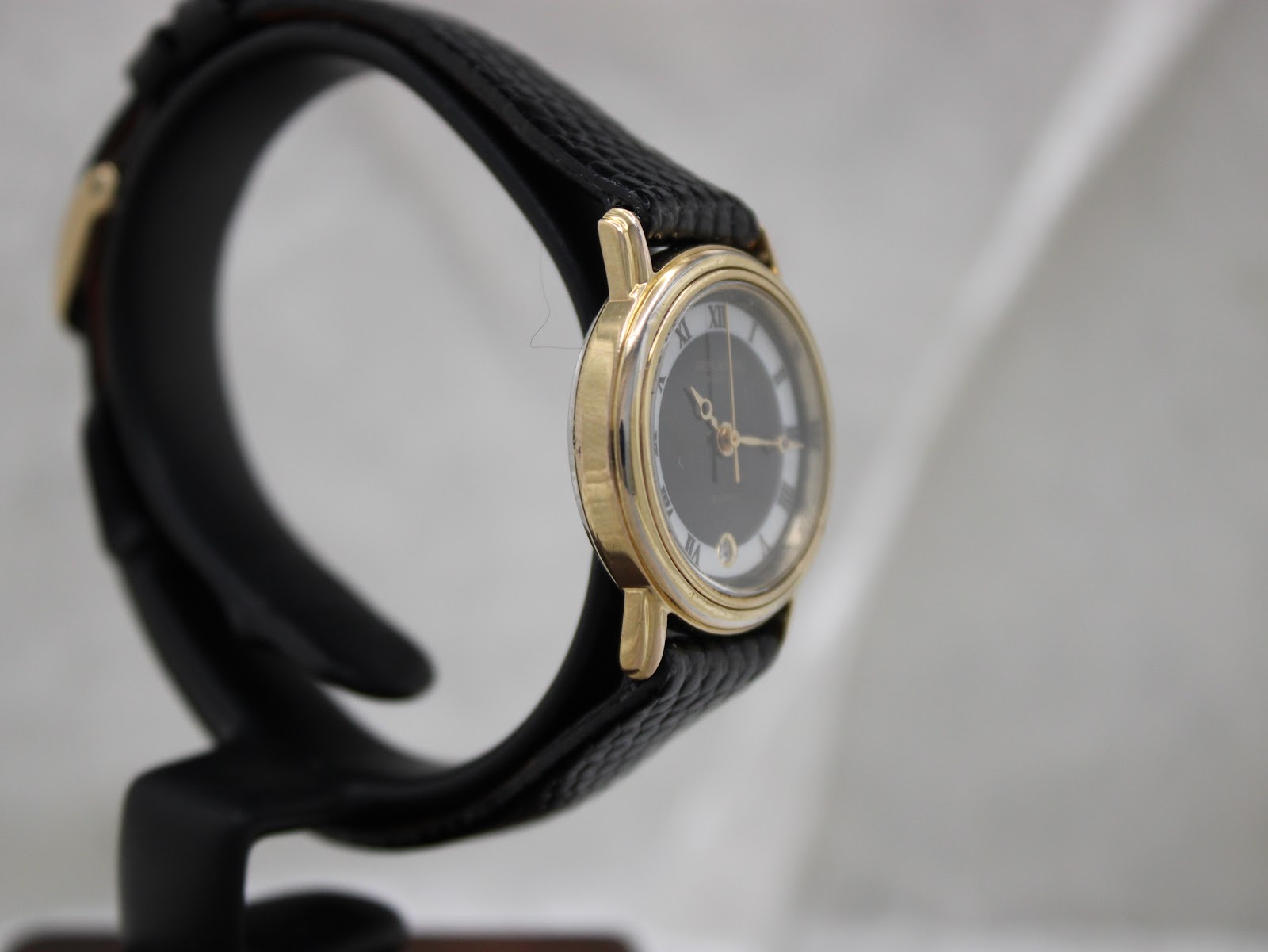Another side profile of the gold-tone Raymond Weil watch, showing the lugs and case side on a black watch stand.