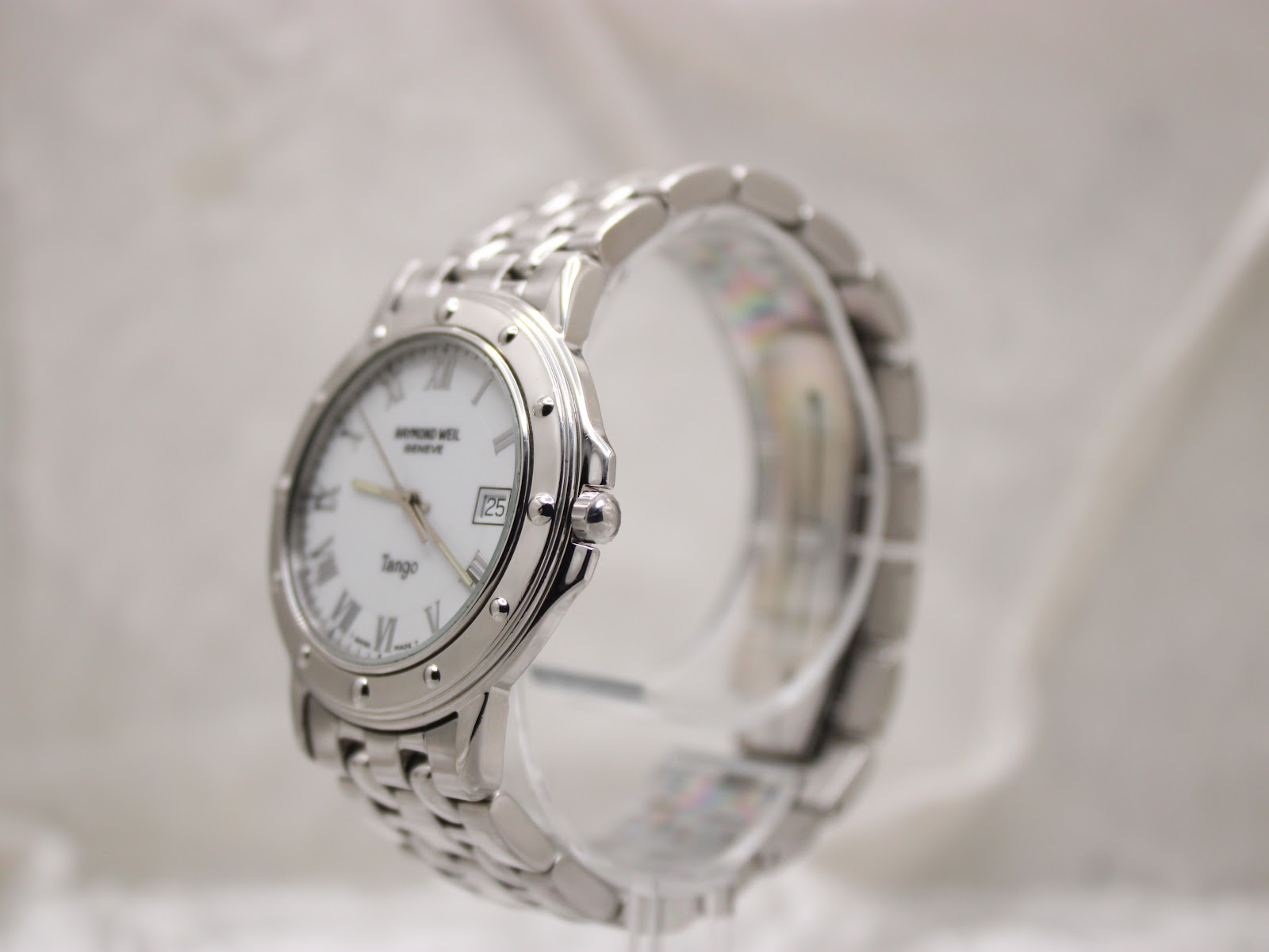 Side view of the Raymond Weil Tango 5560 watch on a clear stand, highlighting the stainless steel case profile and integrated bracelet.