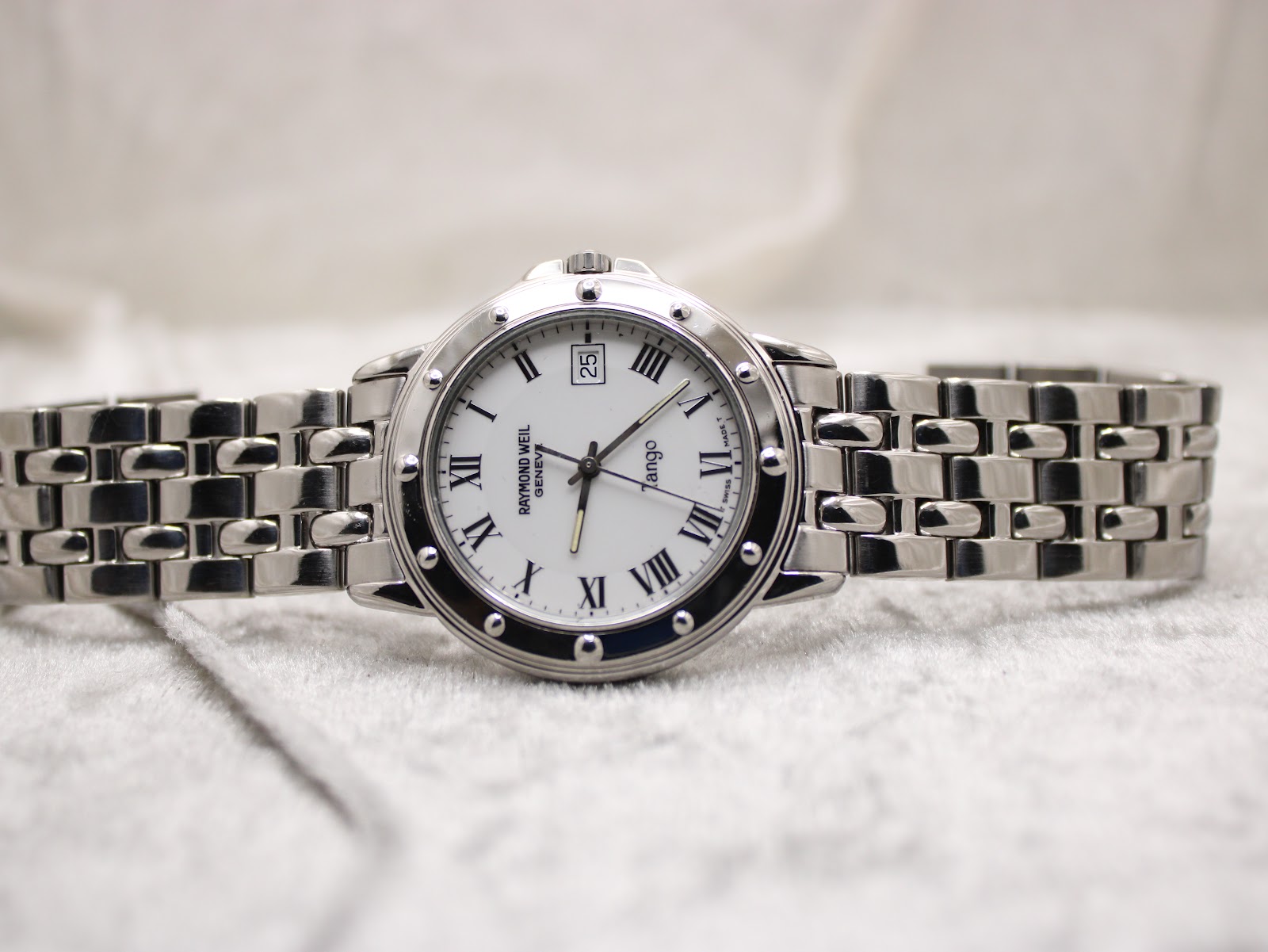 Full front view of the Raymond Weil Tango 5560 watch with its stainless steel bracelet extended on a white textured surface.
