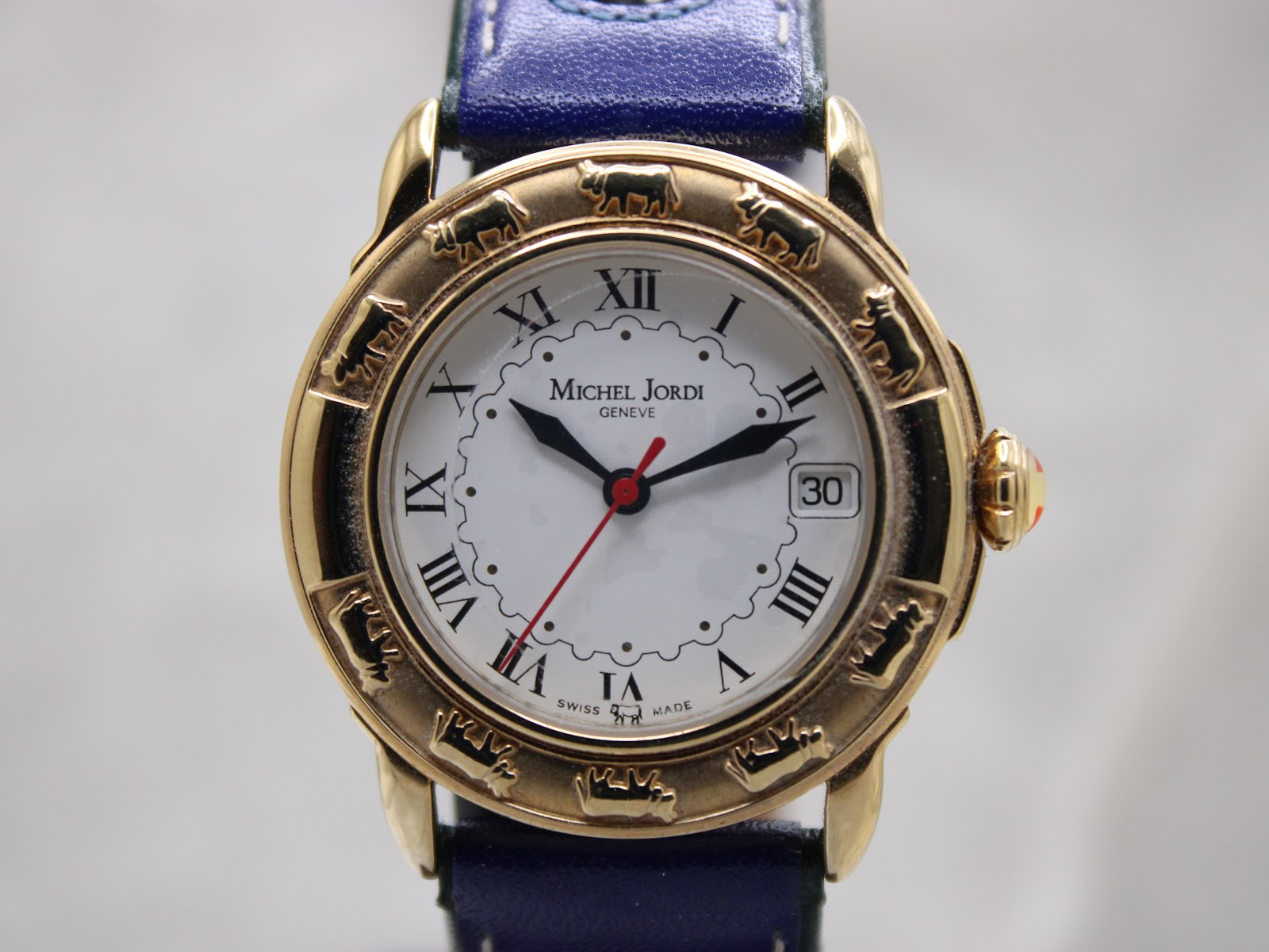 Close-up front view of the Michel Jordi Genève watch dial, showing gold-tone case, Roman numerals, date window, and red seconds hand.