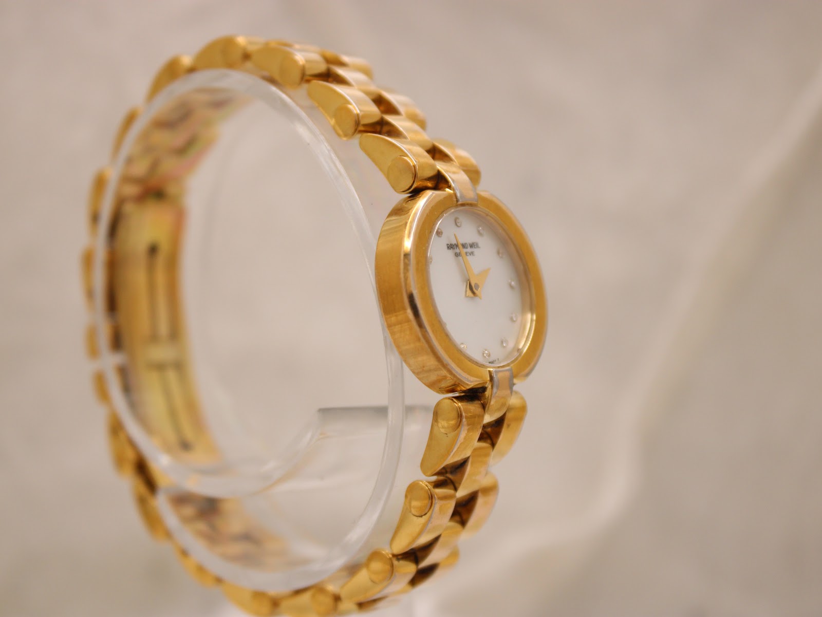 Angled side view of the Raymond Weil watch on a clear stand, highlighting the gold-tone bracelet.