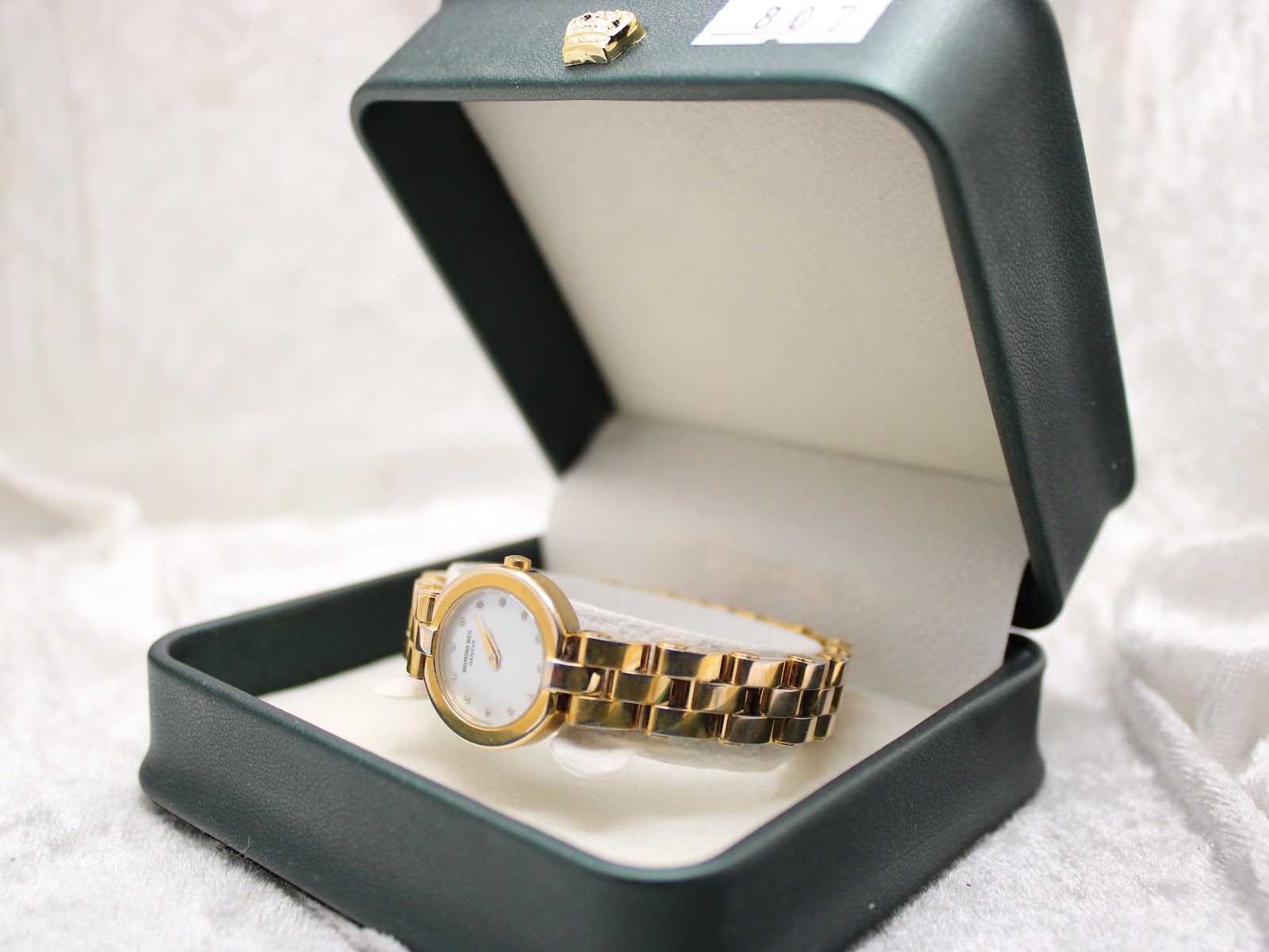 Watch placed inside an open dark green presentation box, showing the dial and gold-tone bracelet.