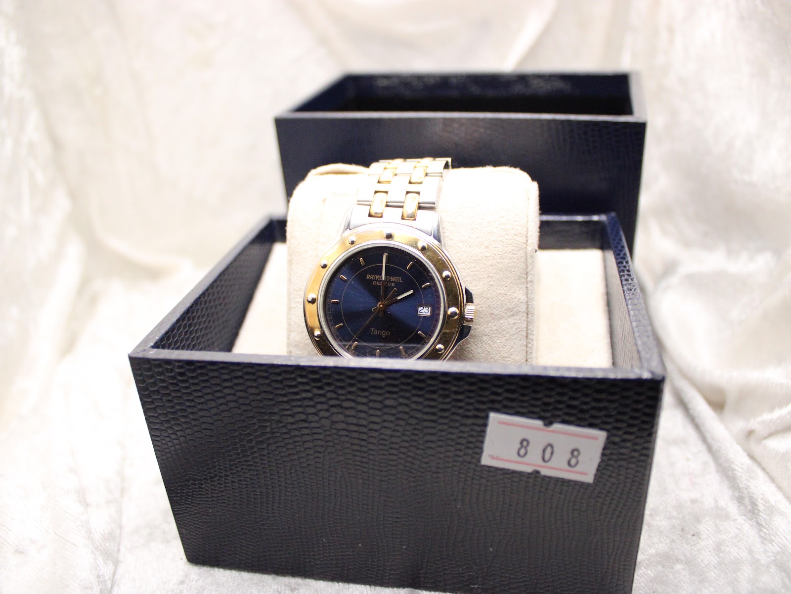 Raymond Weil Tango watch displayed in an open dark blue watch box on a textured surface, with a reference number '808' tag.