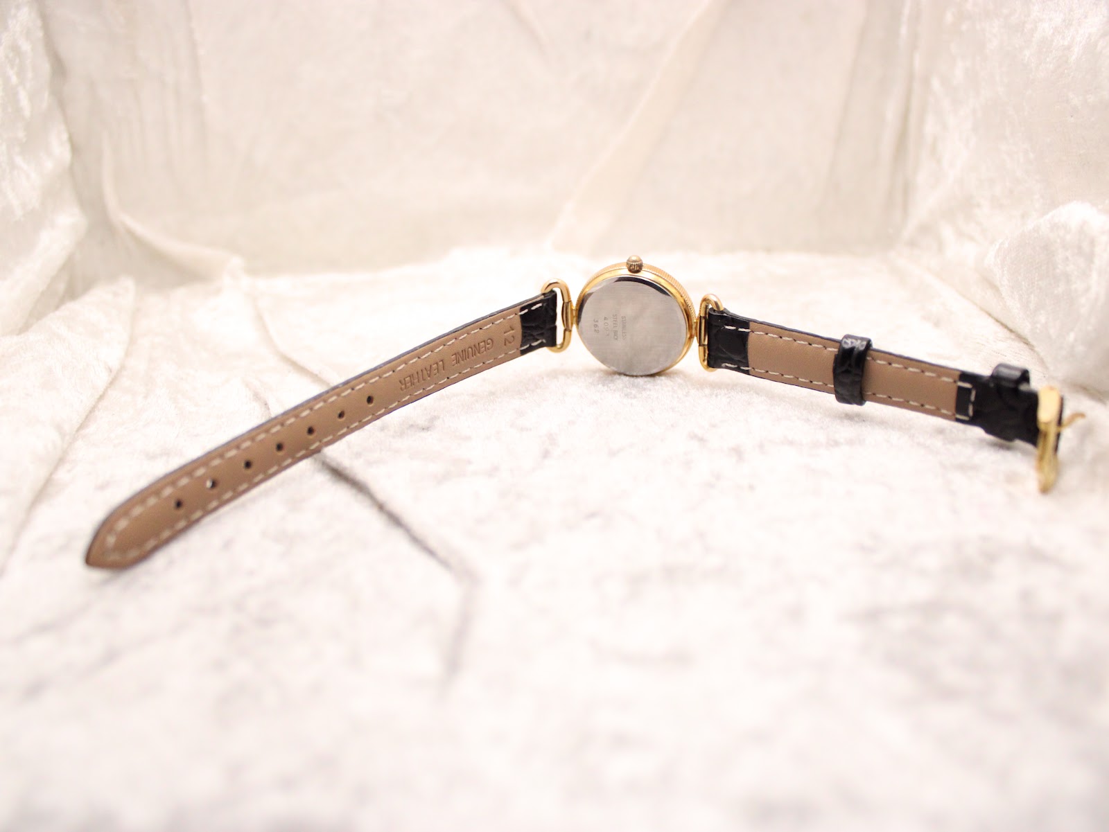 Overhead view of the Rotary watch laid flat on a textured white surface, showing the full length of the black strap and the gold-tone case back.