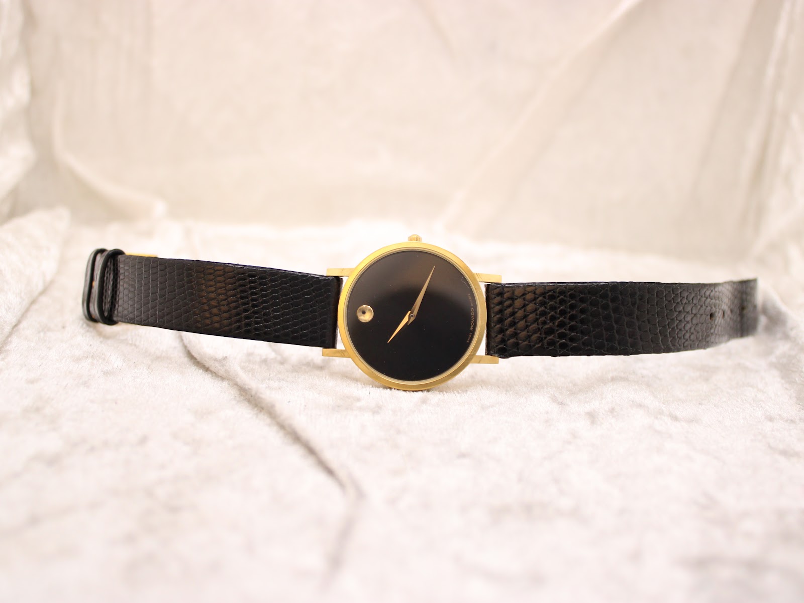 Top-down view of the Movado Museum watch with its black lizard grain leather strap laid flat on a white textured surface.
