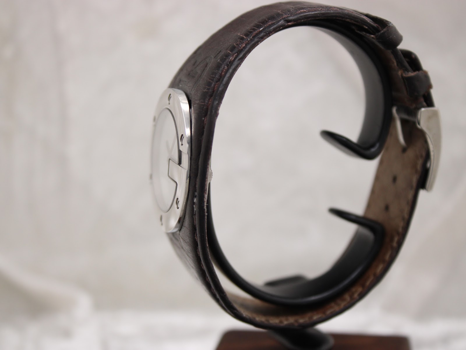 Side view of the Gucci 104 watch on a stand, highlighting the curved profile of the dark brown leather strap and stainless steel case.