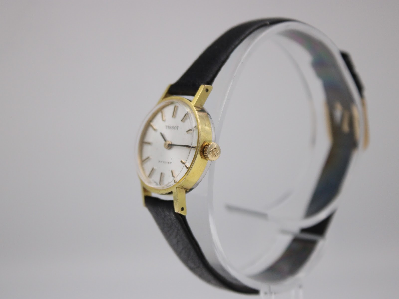 Angled side view of the Tissot Stylist watch showing the gold-plated case, crown, and black strap on a stand.