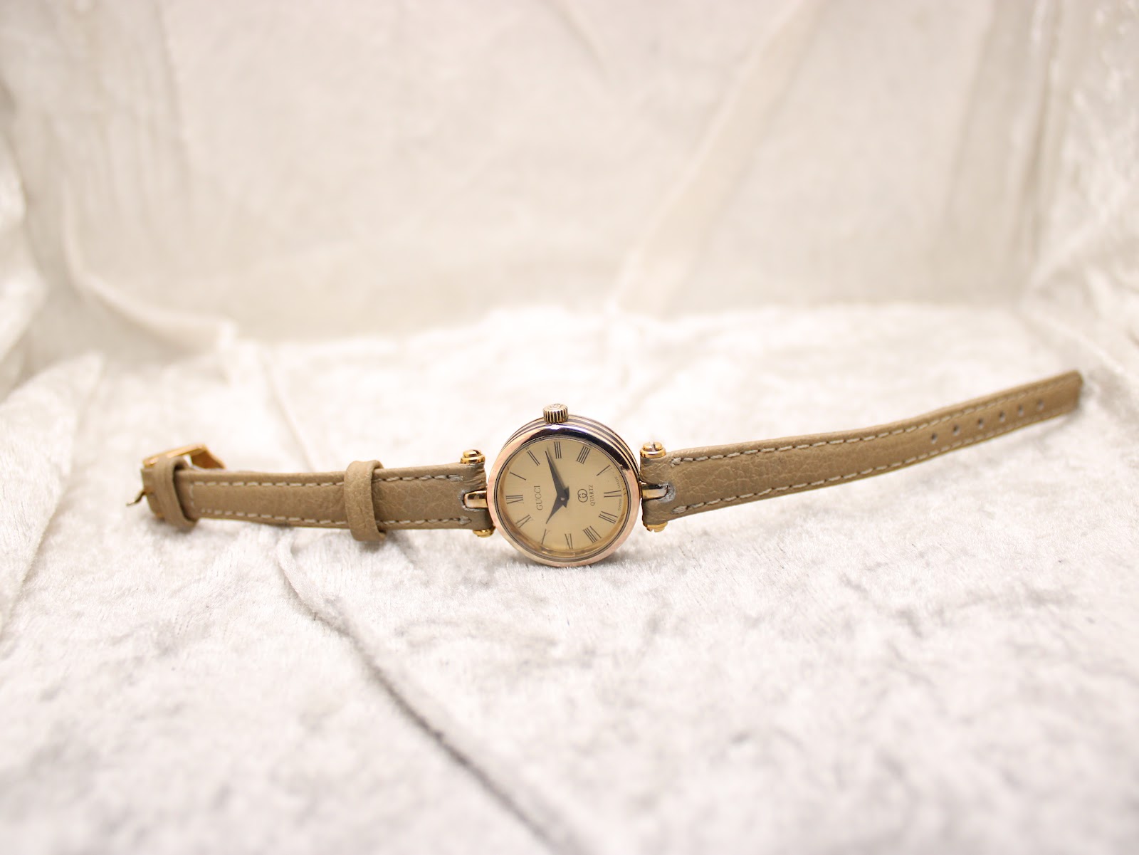 Top-down view of the Gucci 2000L watch with its beige leather strap laid flat on a white textured surface.