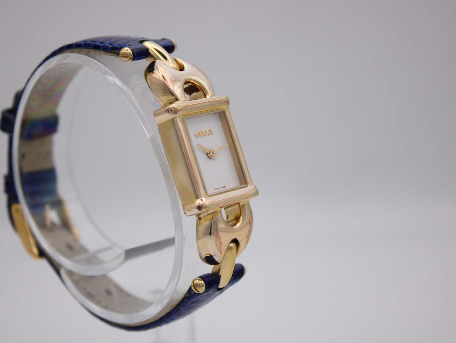 Angled view of the gold plated Gucci 1800 L watch, showing the left side of the rectangular case and blue leather strap.