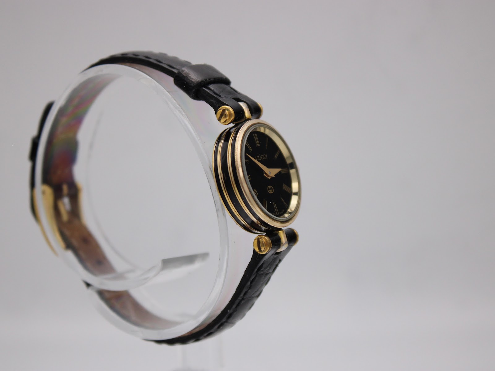 Left side view of the Gucci 2000L watch, showing gold-tone case and black leather strap on a stand.