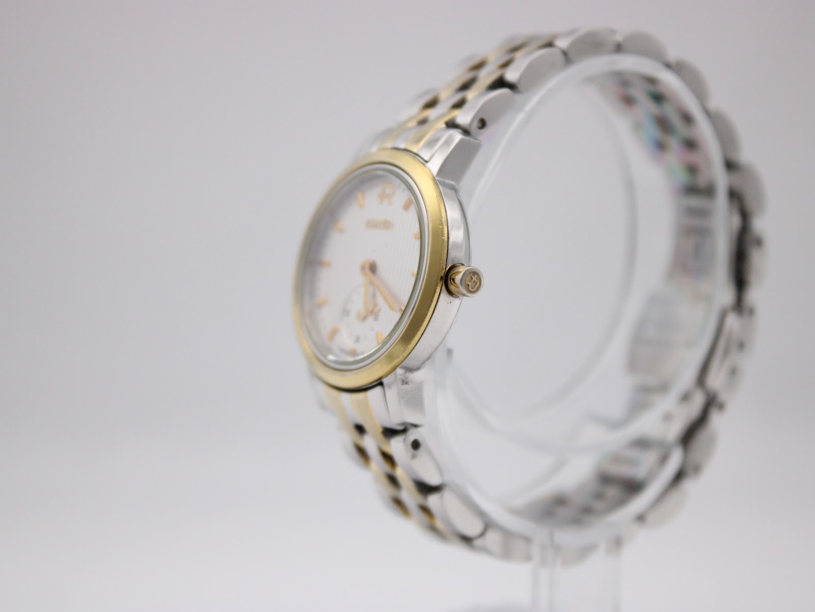 Side-angle view of the Roamer watch on a clear stand, showing the two-tone bracelet and the gold-tone crown with the Roamer 'R' logo.