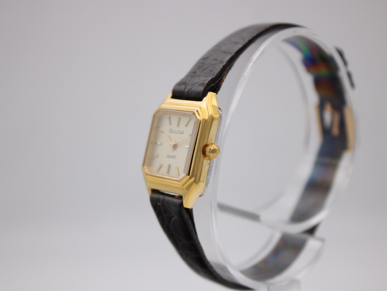 Angled side view of the Bulova watch on a stand, highlighting the gold-tone case and crown, with the dark brown leather strap.