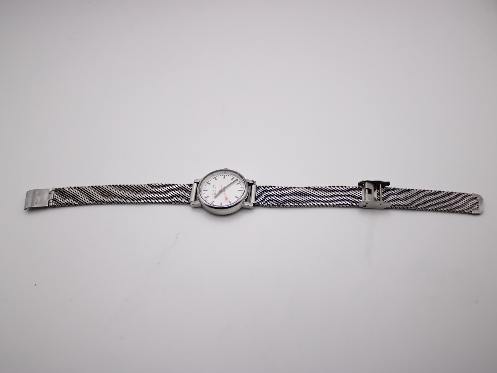 Top-down full length view of the Mondaine watch and open mesh bracelet on a white background.