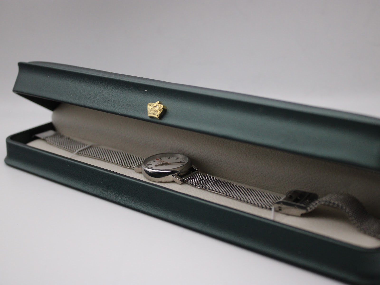 Mondaine watch with mesh bracelet displayed inside a dark green presentation box.