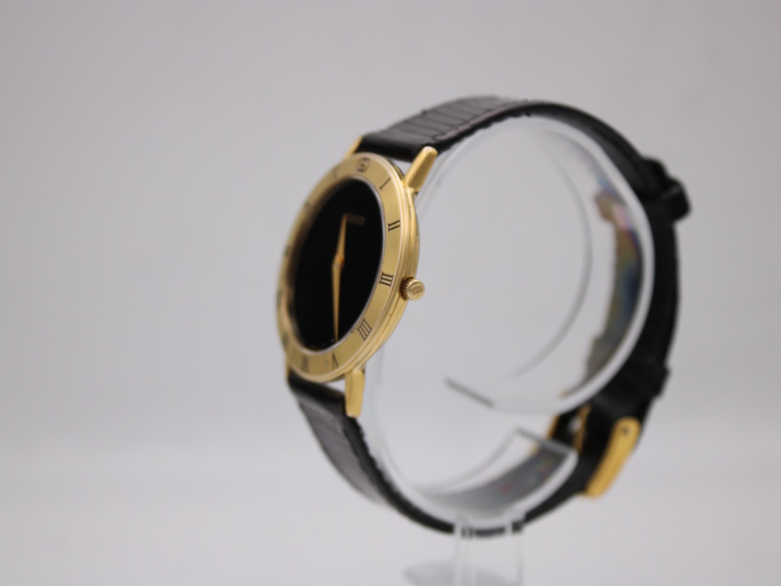 Angled side view of the Gucci 3000.2.M watch, showcasing its slim gold-tone case and black leather strap on a clear display stand.