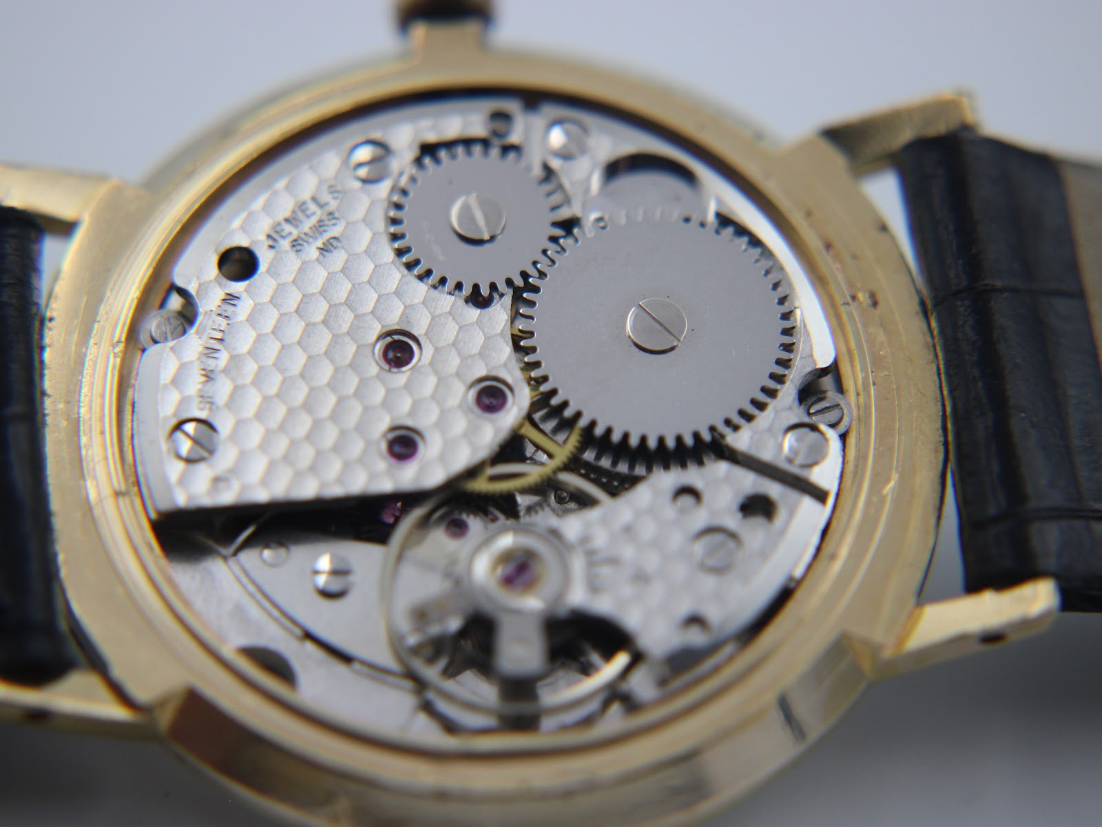 Detailed view of the Swiss Made manual wind mechanical movement with various gears, jewels, and the balance wheel visible.