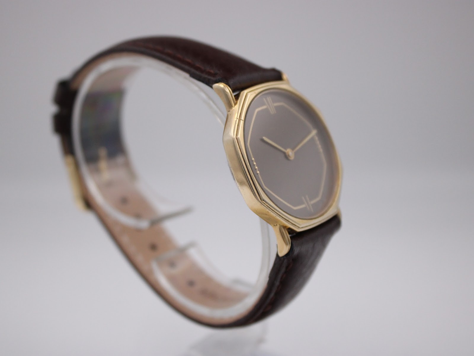 Another angled side view of the watch on a stand, showing the curved brown leather strap and the slim profile of the gold case.