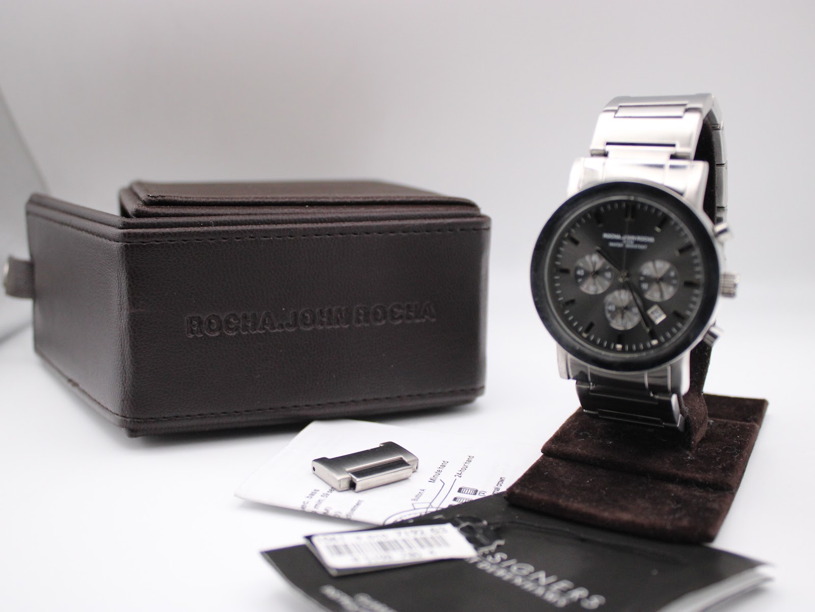 Watch on display stand with its original Rocha John Rocha presentation box, booklet, and spare bracelet link on a white background.