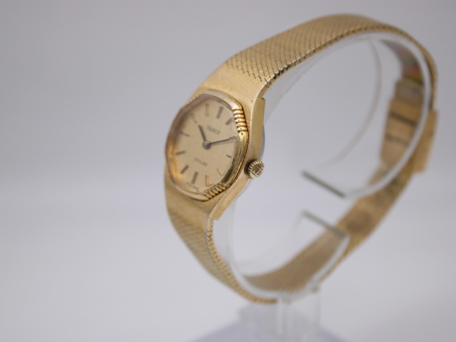 Angled view of the gold Tissot Stylist showing the crown and integrated bracelet on a clear stand.