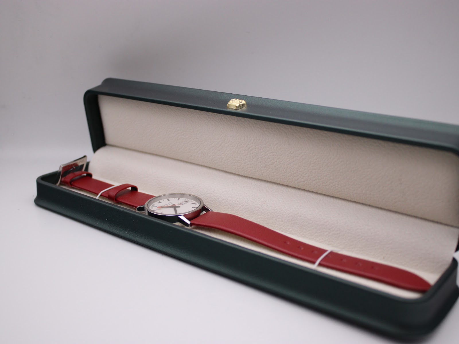 Mondaine watch with red strap presented inside a dark green, cream-lined watch box, ready for presentation.