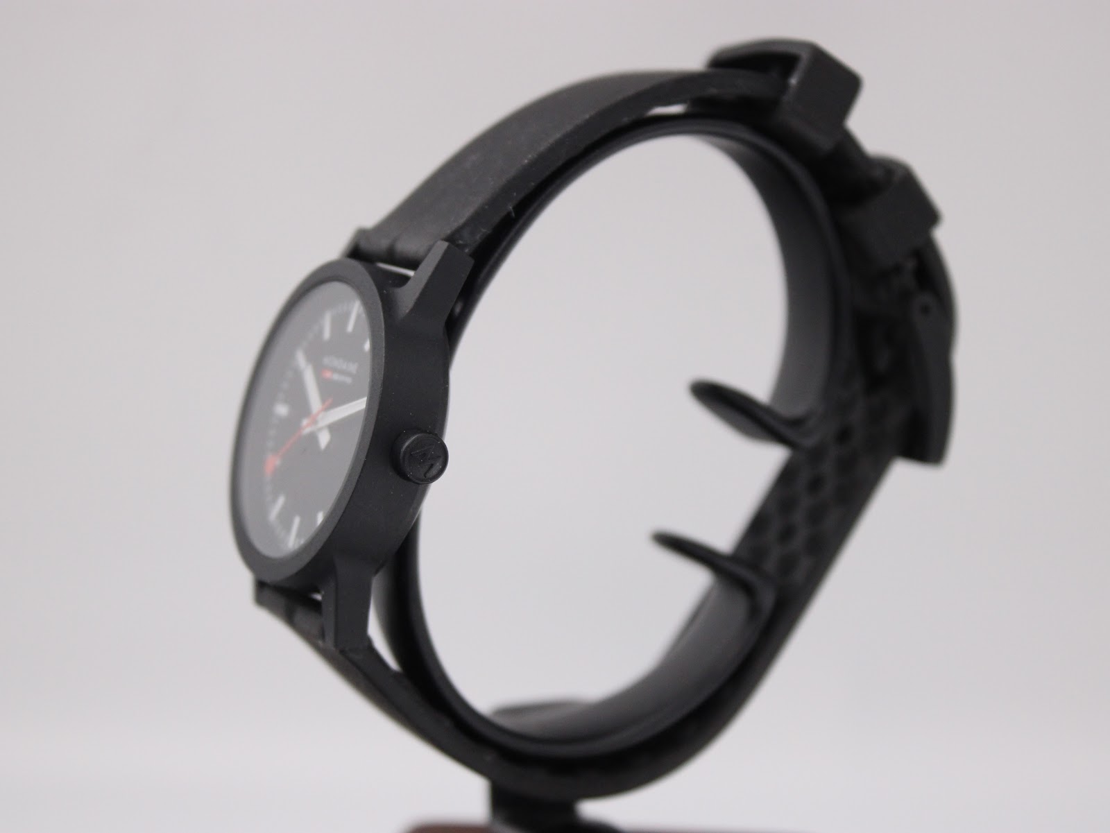 Side view of the black Mondaine watch on a display stand, showing the case profile and rubber strap