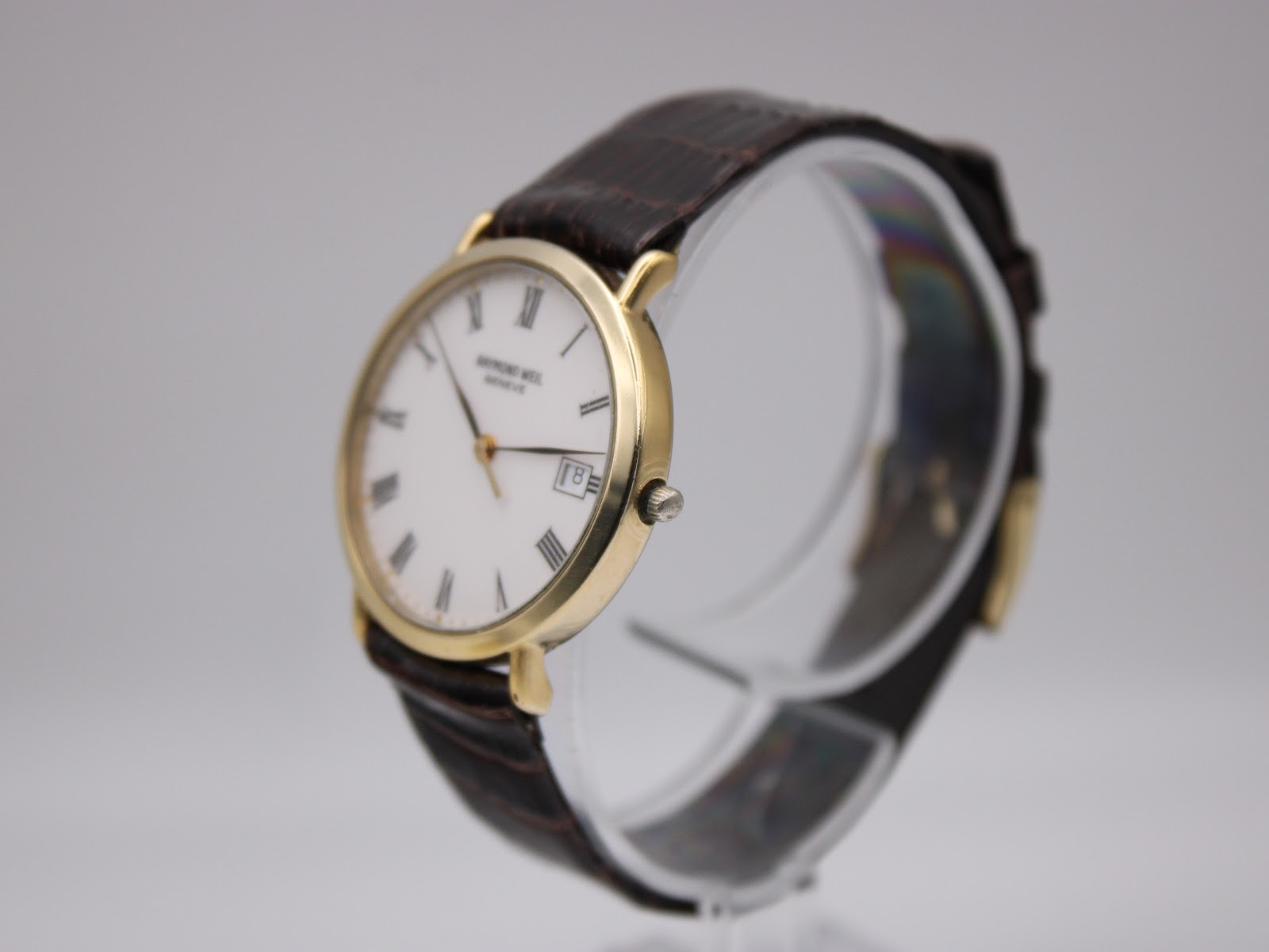 Angled view of the Raymond Weil dress watch on a clear display stand, showing the right side of the gold case and crown.