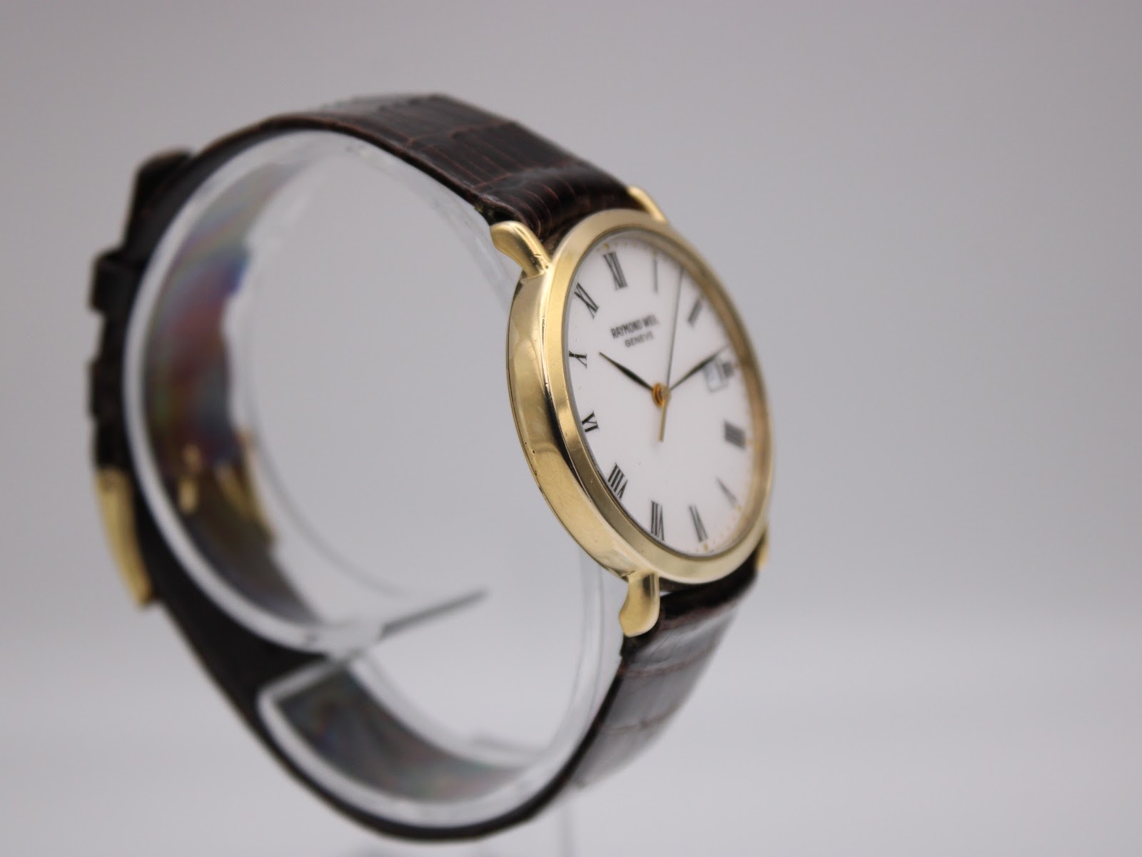 Side view of the Raymond Weil Geneve watch on a clear display stand, showing the gold case and brown leather strap.