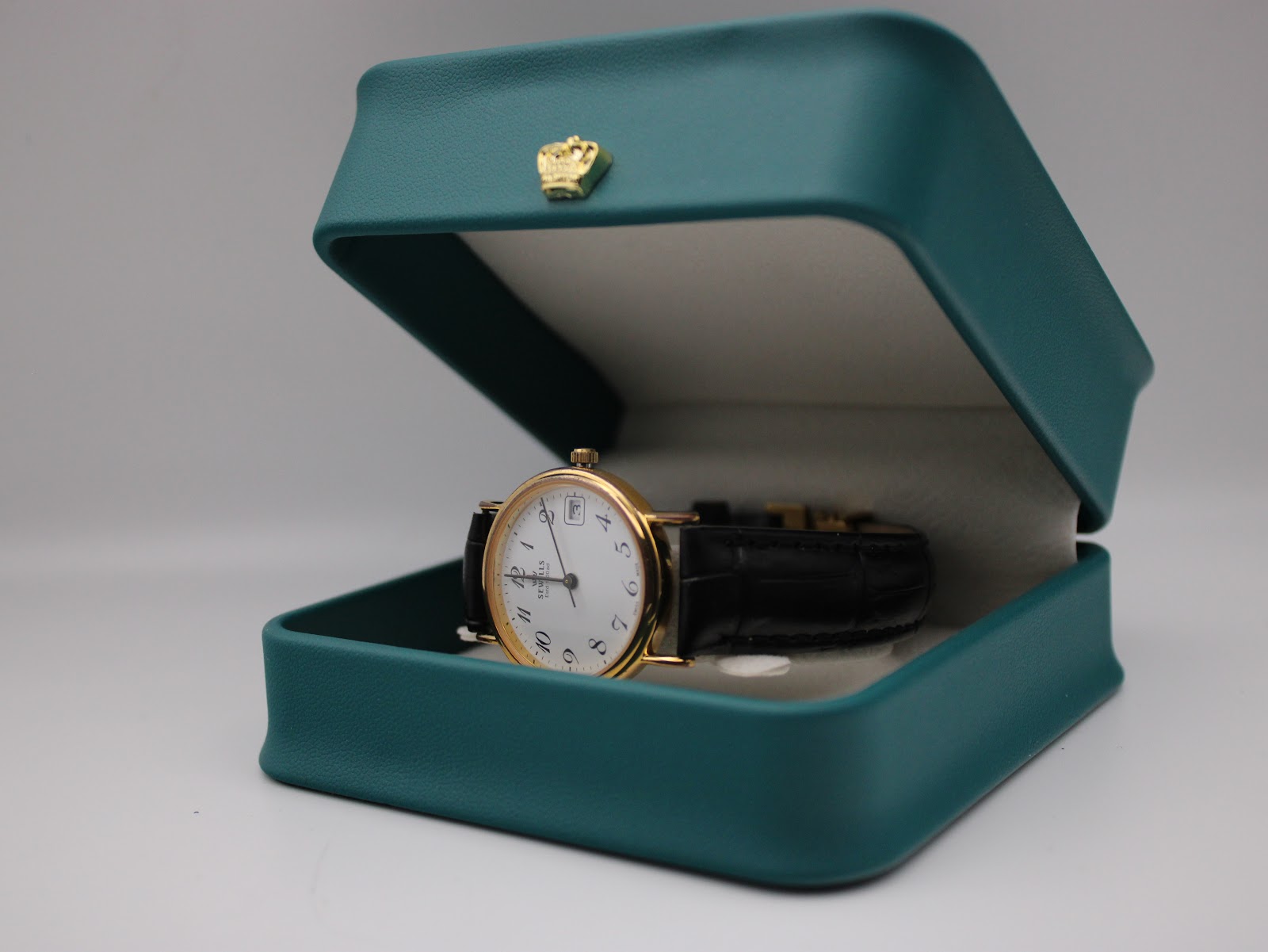 The Sewills watch positioned inside an open teal presentation box with a small gold crown emblem.
