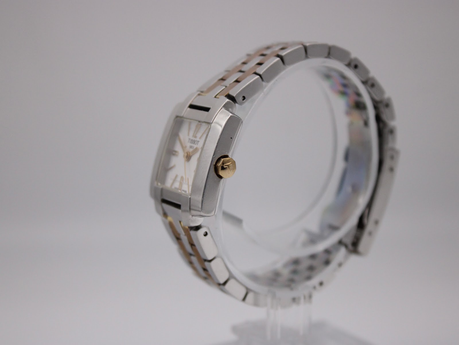 Angled side view of the Tissot T-Trend watch on a clear display stand, highlighting the profile of the case and the gold-tone crown.