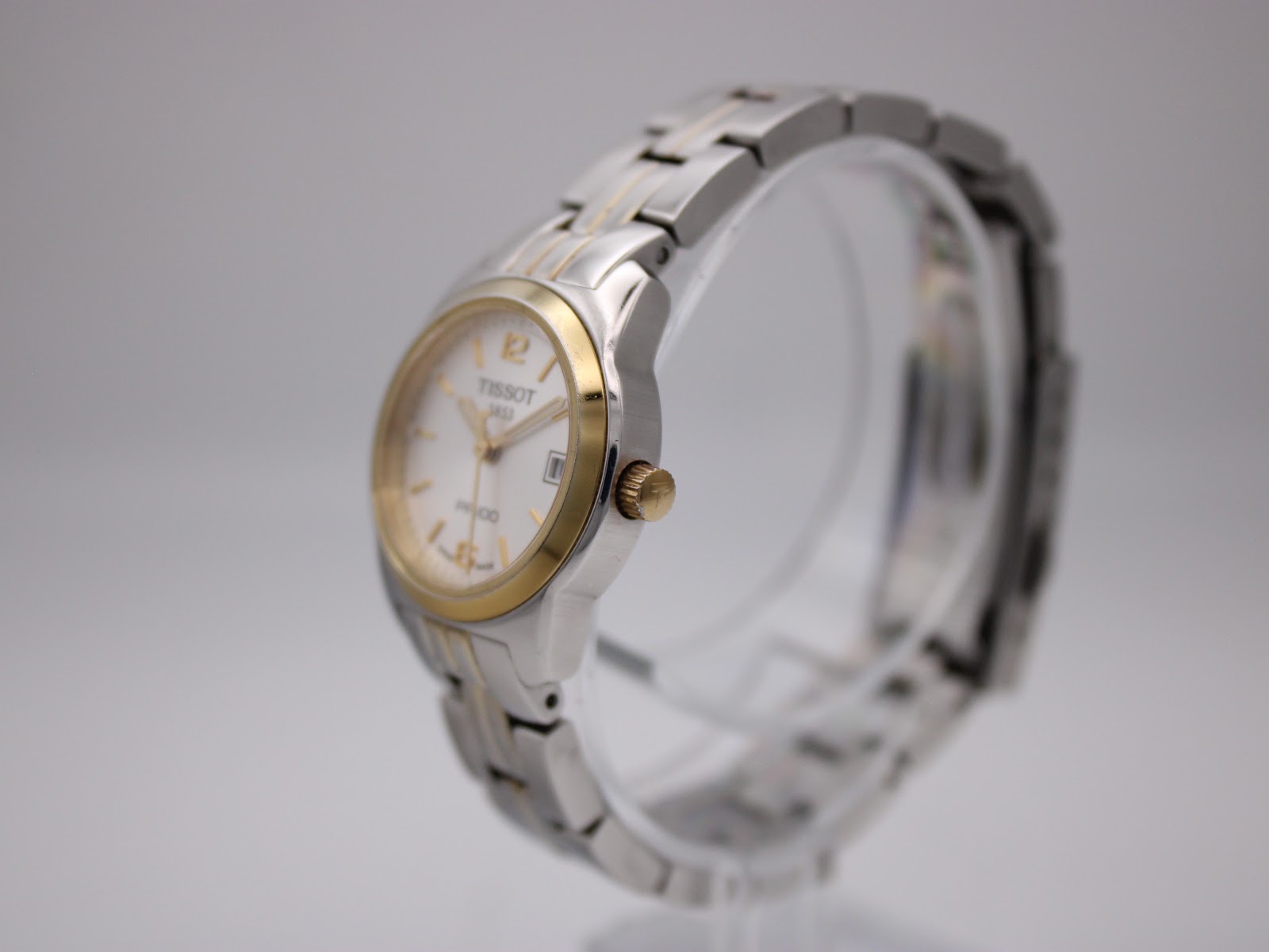 Angled side view of the Tissot watch on a clear display stand, showing the curved case and bracelet links.
