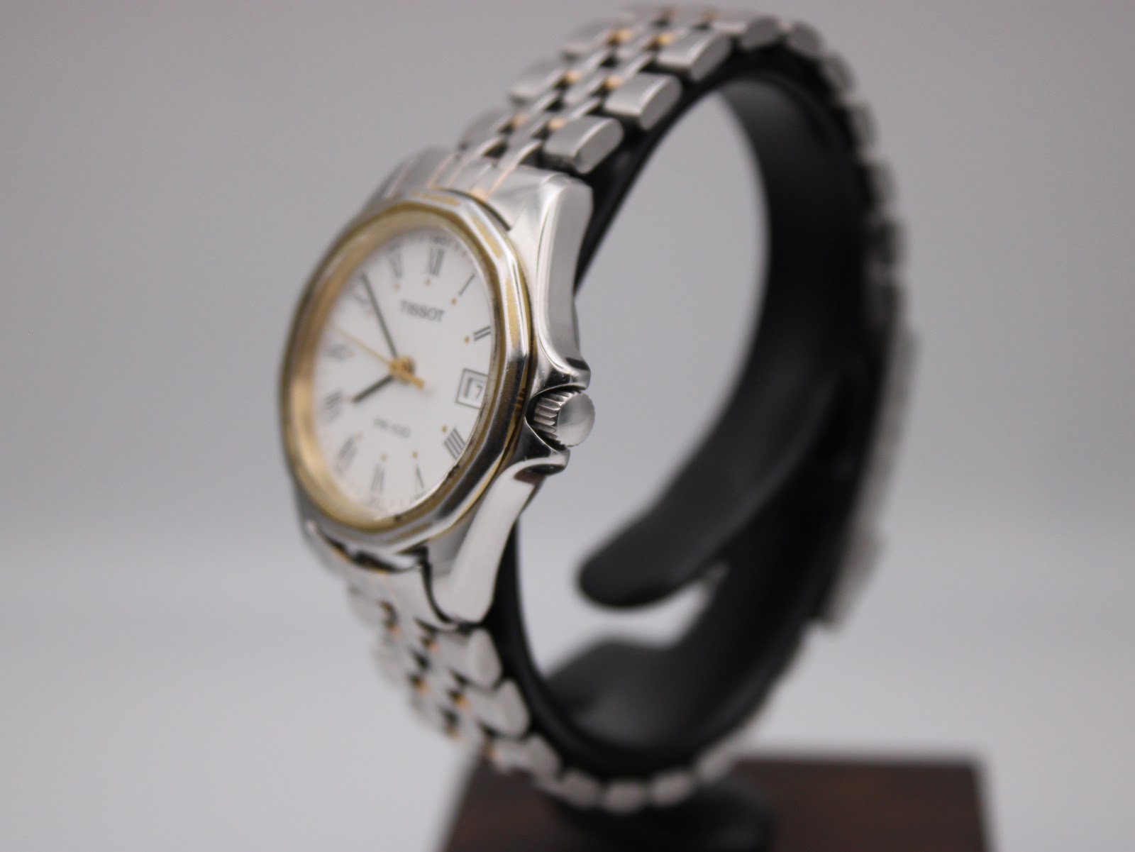 Angled side view of the Tissot watch from the right, highlighting the crown and polished case profile, with the two-tone bracelet curving around a display stand.