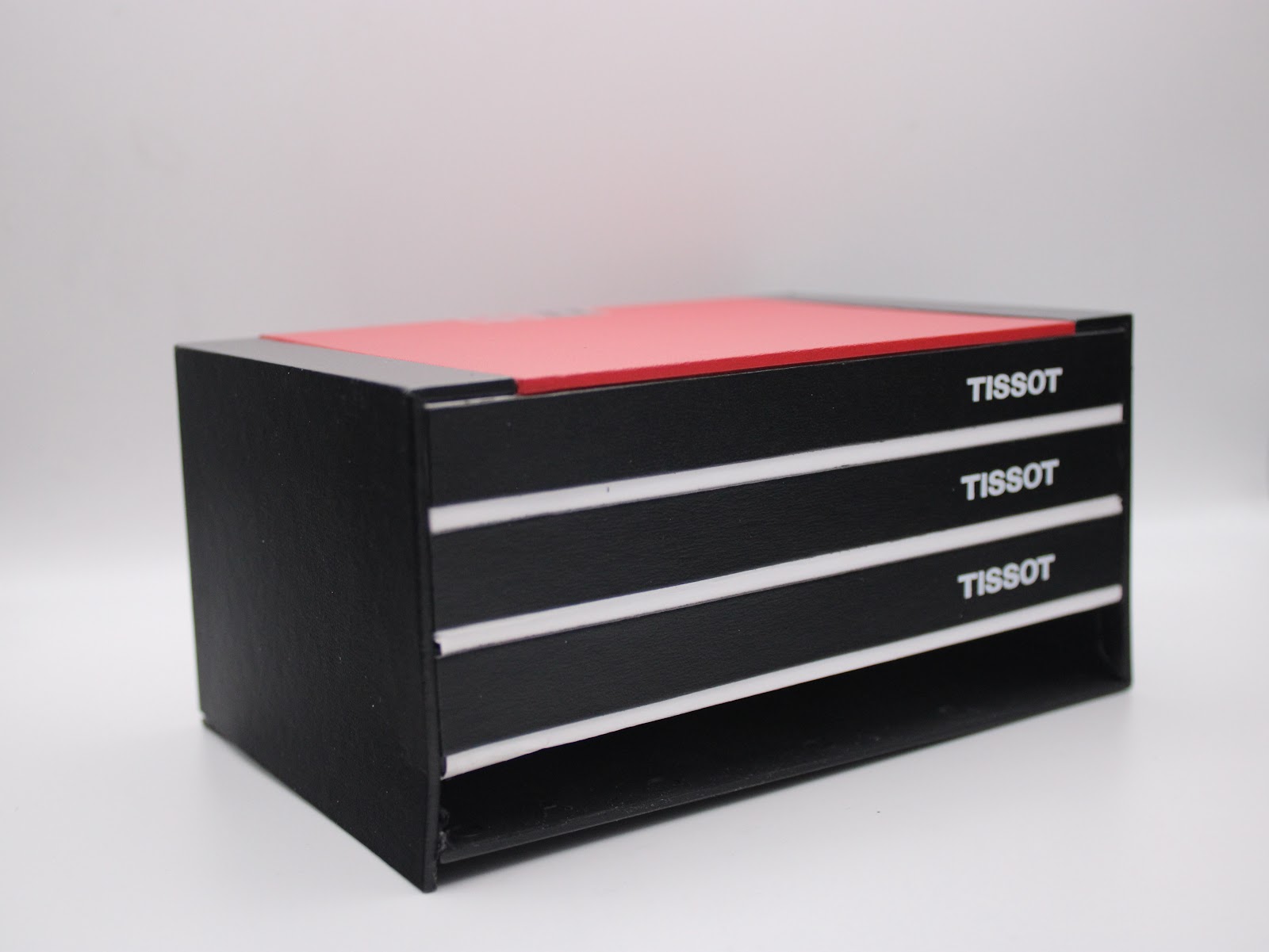 Angled view of the Tissot original presentation box, black with a red top and white 'Tissot' branding on the drawers.